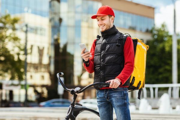 Courier Using Cellphone Delivering Food From Restaurants On Bicycle Outdoors