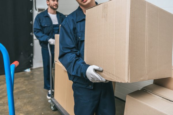 cropped view of two movers in uniform transporting cardboard boxes with hand truck in warehouse